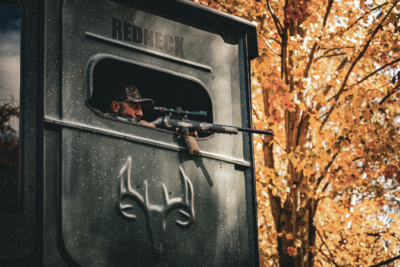 Hunter aiming a Weatherby Model 307 Alpine CT rifle from a Redneck hunting blind surrounded by vibrant autumn foliage.